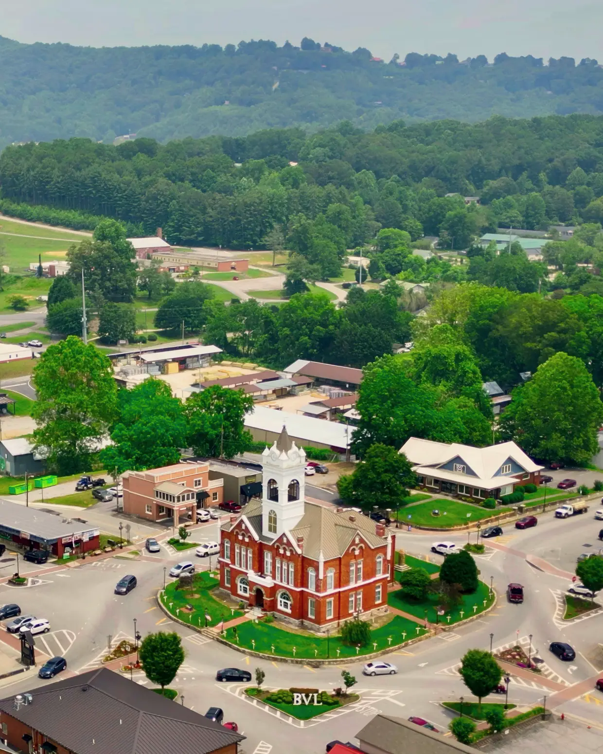 Vertical drone view of the Old Union County Courthouse on its round green square, with surrounding downtown roofs, trees and a wooded ridge on the horizon.