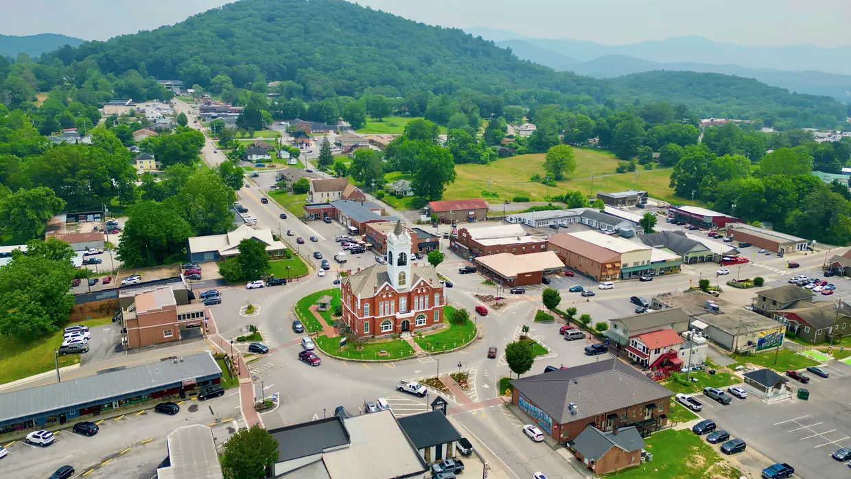 Drone view over downtown Blairsville with the red-brick 1899 Old Union County Courthouse and its white clock tower on the green courthouse square, ringed by roads, shops and parking, with forested mountains behind.