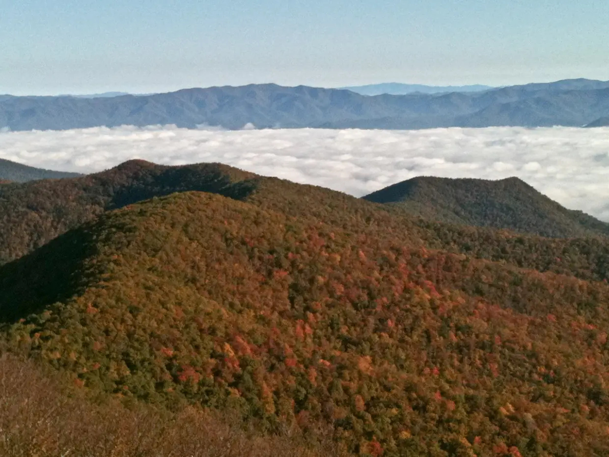 Early-morning view north over orange and green fall-colored peaks, with fog filling the valley and distant blue ridges above it.