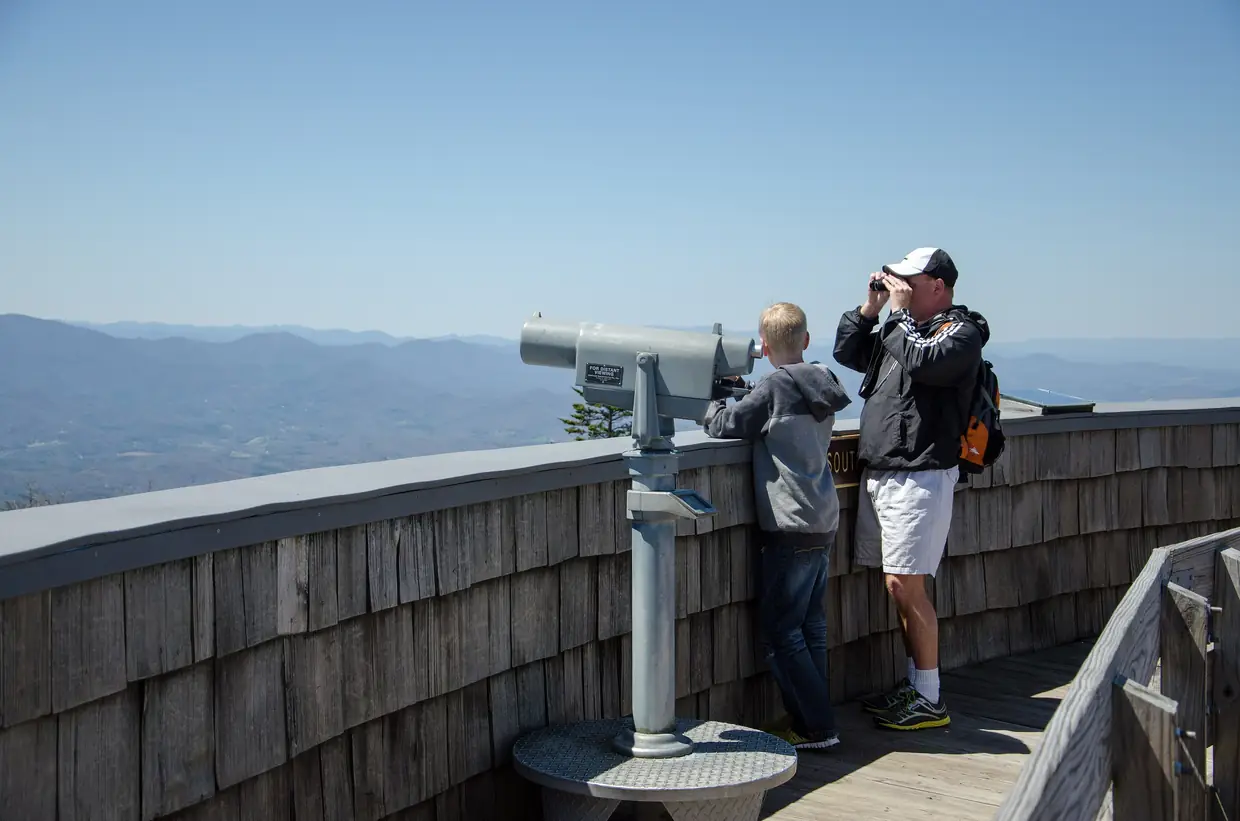 Two visitors, an adult and a boy, look out with binoculars and a coin viewer from the shingled wall of the summit observation deck over hazy blue mountains.