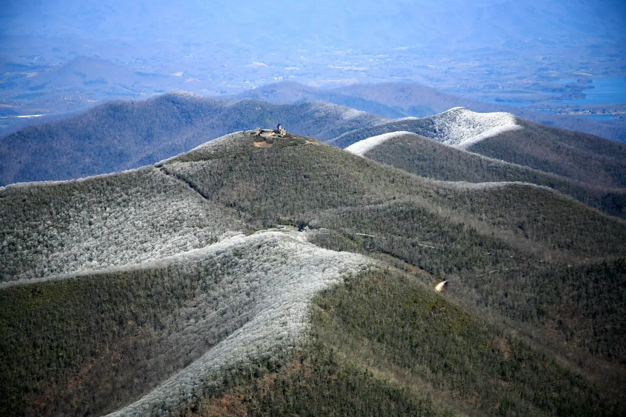 Aerial view of Brasstown Bald's summit and visitor center on a ridge dusted white with rime ice, with layered ridges, valleys and a lake in the hazy distance.