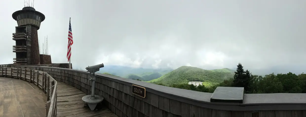 Wide panorama from the summit observation deck, with the stone lookout tower and flag at left, a coin-operated viewer, and green ridges under low clouds.