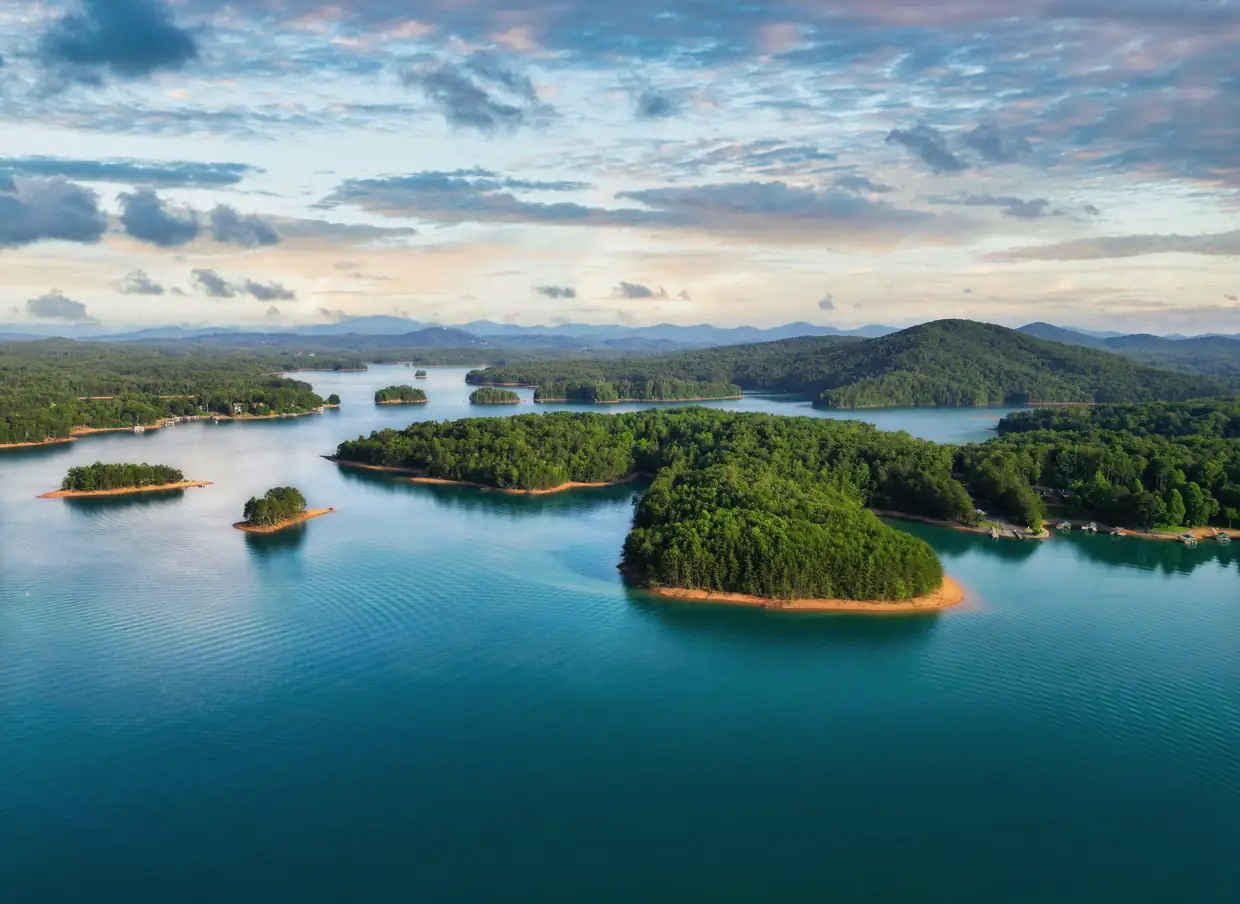 Drone view of Lake Nottely's turquoise water with wooded islands and red-clay shorelines, under a pastel sky with scattered clouds and mountains on the horizon.