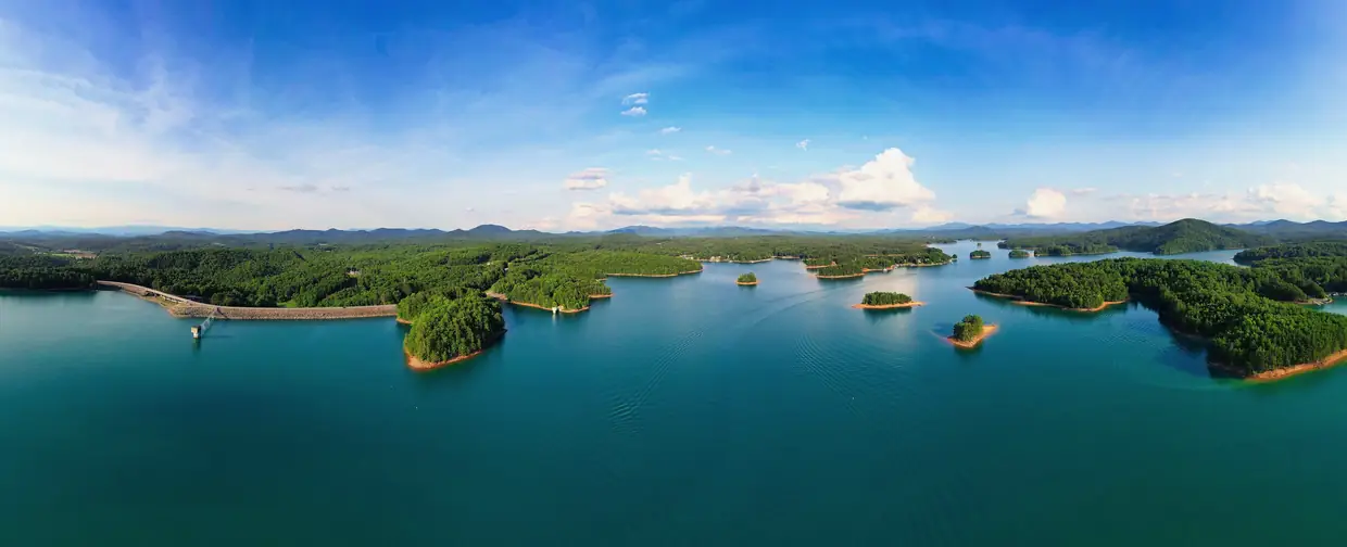 Wide aerial panorama of Lake Nottely's teal water, wooded islands and shoreline, with the dam at far left and blue sky with white clouds.