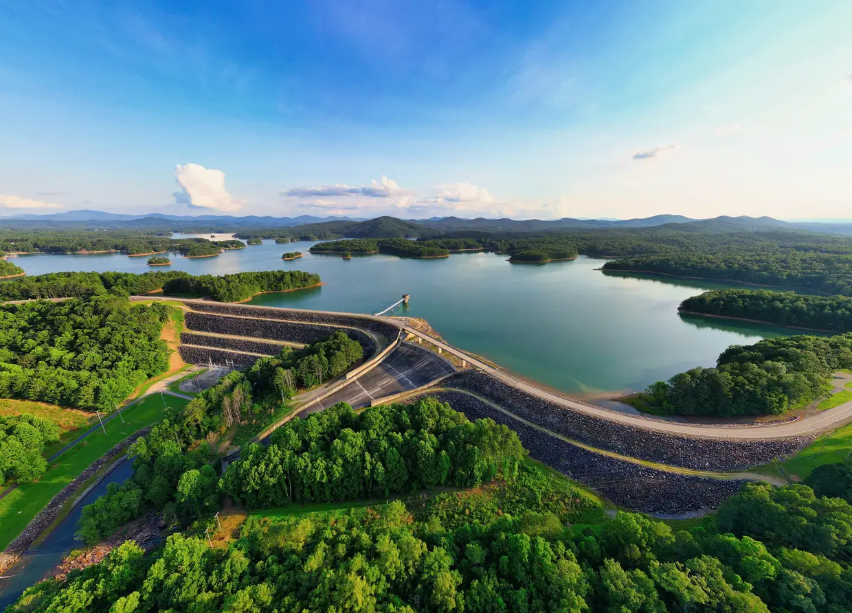 Aerial view of the curving rock-faced Nottely Dam with a road across its crest, Lake Nottely stretching beyond, and green forest in the foreground.