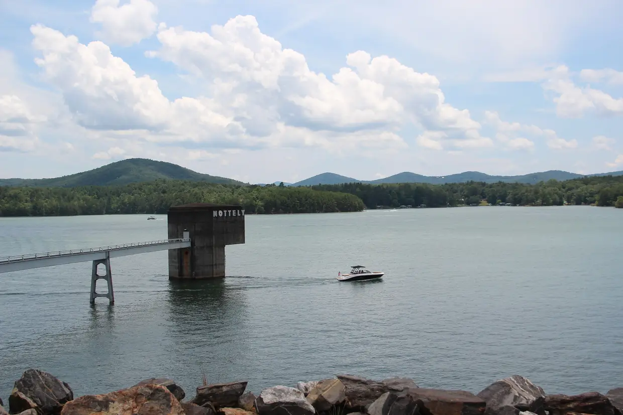 From the rocky shore, the concrete intake tower labeled NOTTELY stands in the lake on a footbridge while a motorboat passes, with wooded mountains behind.