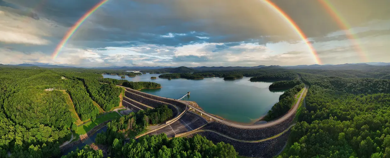 Wide aerial panorama of Nottely Dam curving across the lake under a double rainbow, surrounded by green forested hills.