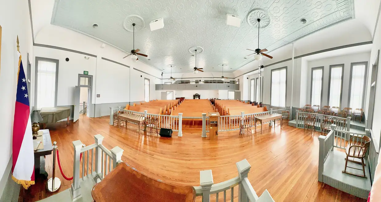 Wide panoramic interior of the restored courtroom, with wooden pews, a railed judge's area, pressed-tin ceiling, ceiling fans, tall windows and a Georgia flag.