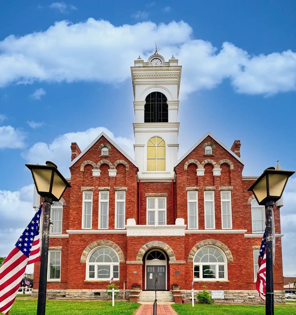 Front facade of the red-brick Old Union County Courthouse with arched windows and a white bell tower, framed by two black lamp posts and American flags under a bright cloudy sky (heavily color-processed).