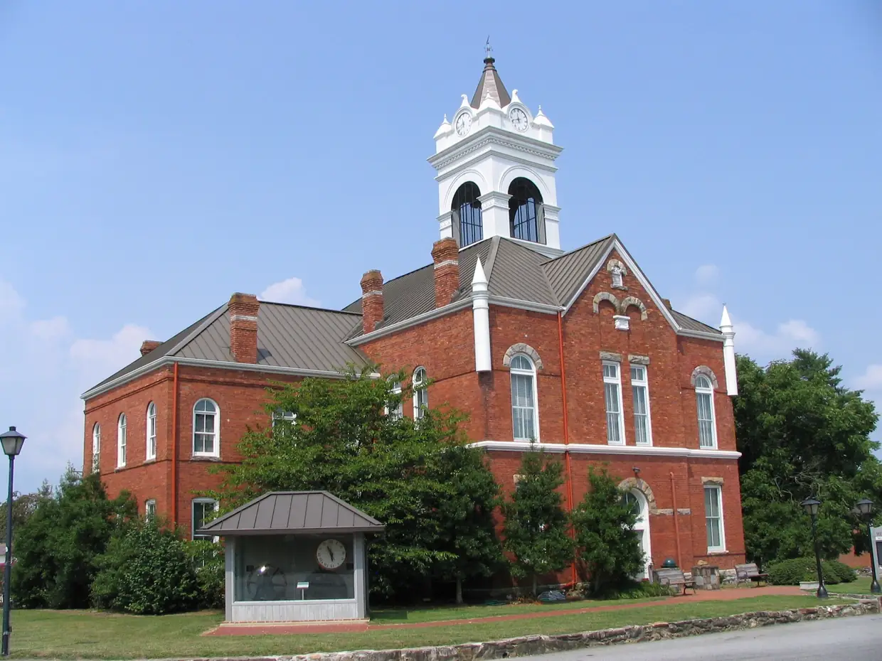 Side view of the red-brick courthouse and white cupola behind shrubs and trees on the lawn, with a small roofed kiosk in front, under a clear blue sky.