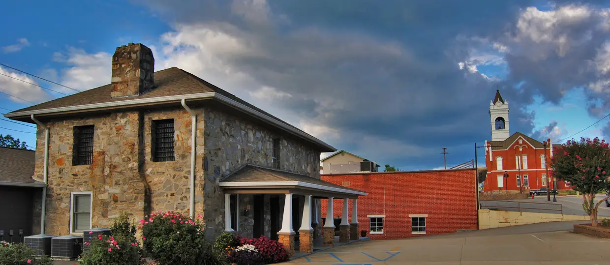 The stone 1934 Old Union County Jail with its columned porch in the foreground and the courthouse's white tower and red brick visible down the street under dramatic clouds.