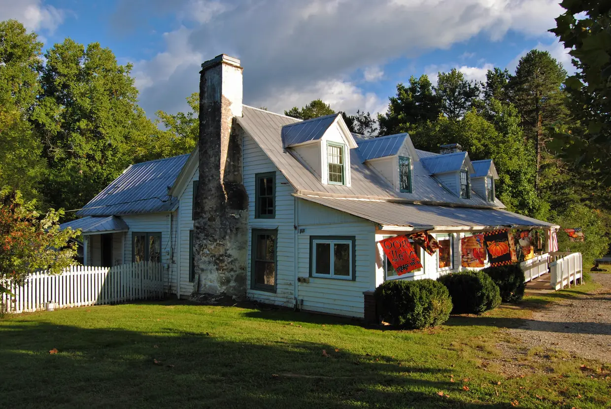 A white 1885 farmhouse with a blue metal roof, dormers, a tall stone chimney and a picket fence, with colorful rugs on the porch rail.