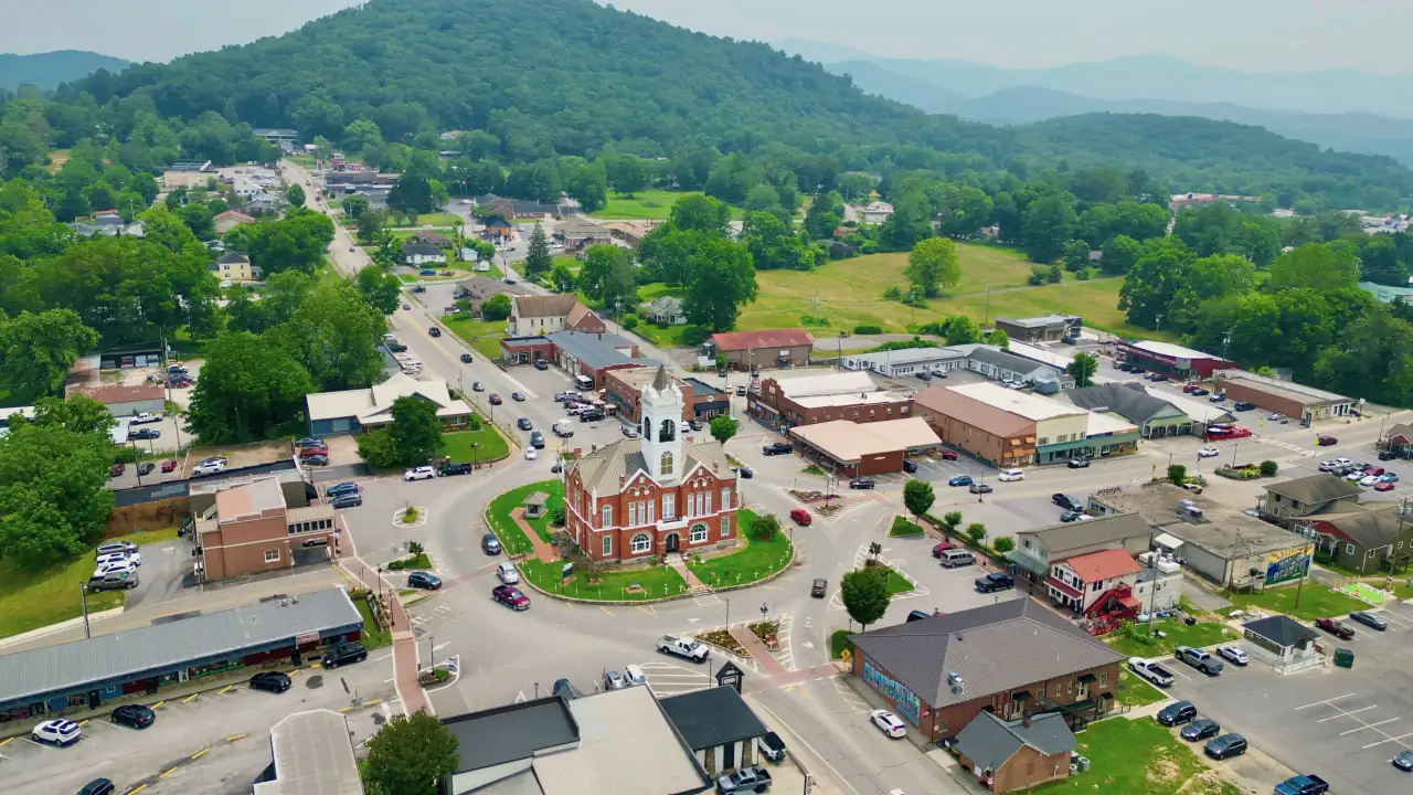 Drone view over downtown Blairsville with the red-brick 1899 Old Union County Courthouse and its white clock tower on the green courthouse square, ringed by roads, shops and parking, with forested mountains behind.