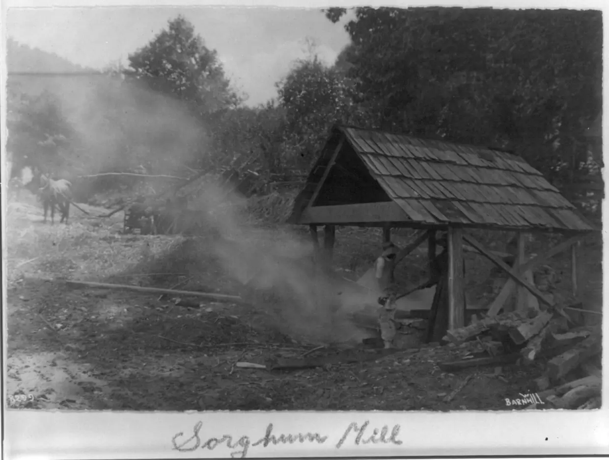 Old black-and-white photo of a shingle-roofed sorghum boiling shed with steam rising, a horse-powered cane mill in the background and the handwritten caption 'Sorghum Mill'.