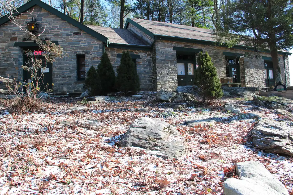 The low stone Walasi-Yi Interpretive Center with green doors and trim, small evergreens and rocks in front, and fallen leaves and light snow on the ground.